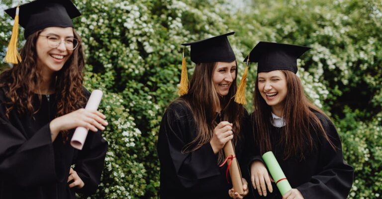 Anel de formatura é obrigatório? Entenda a tradição e as escolhas atuais Três mulheres em trajes de formatura sorrindo e segurando diplomas em um ambiente natural.