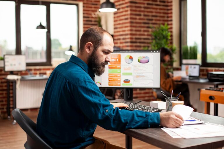 Homem de barba e camisa azul confere papéis na mesa diante de um monitor com gráficos de desempenho em escritório